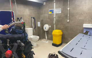 A changing places toilet facility with a large changing bench, hoist, toilet and bin visible in the photograph. The walls are tiled grey and the floor is tiled white. There is a black and white handbag in the middle of the floor. A teenage boy with complex needs is sitting in his black wheelchair with his back to the camera.