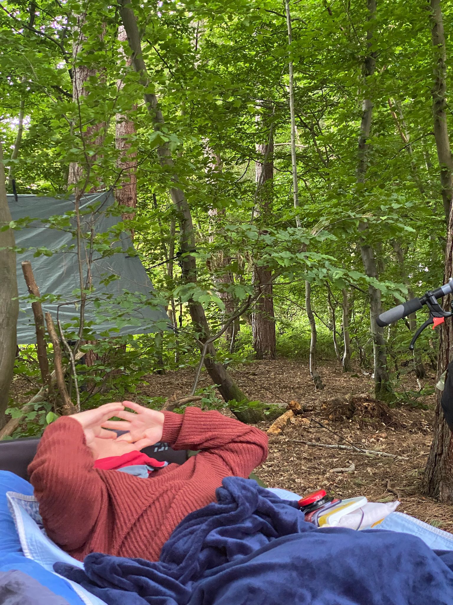 Young man with complex needs wearing a rust coloured jumper and smuggled in a blue velour blanket is lying on a camp bed outdoors in a clearing.