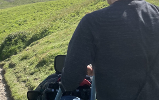 Father and son are walking together in the countryside. Dad is wearing a black top and jeans, pushing his son in a wheelchair along a stoney trail.