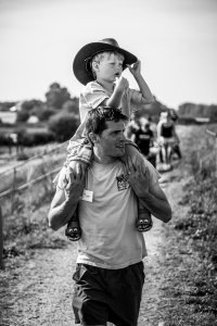 A young boy (wearing his Dad's cowboy hat) sitting on his dad's shoulders walking along a seawall.